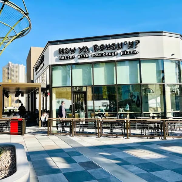 The exterior of How Ya Dough'n Pizza, with outdoor seating and a modern glass facade.