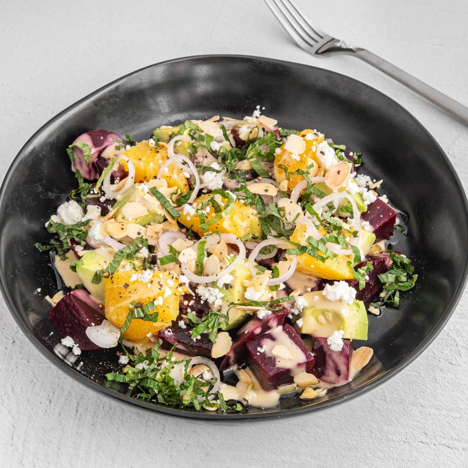 Beet and orange salad with avocado, feta, almonds, and herbs in a black bowl.