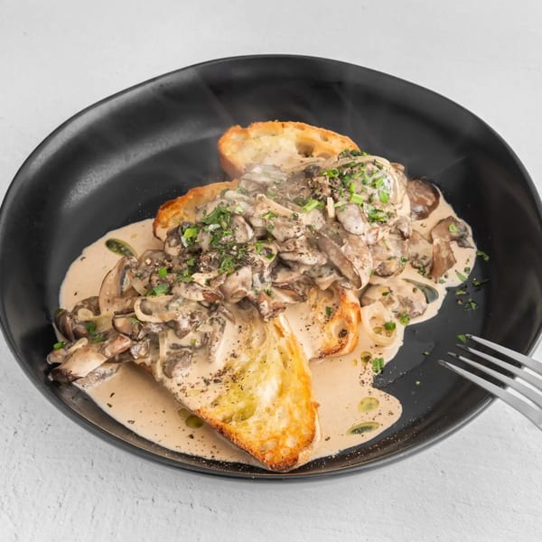 Steaming creamy mushroom toast with chives and pepper on a black plate with a fork.