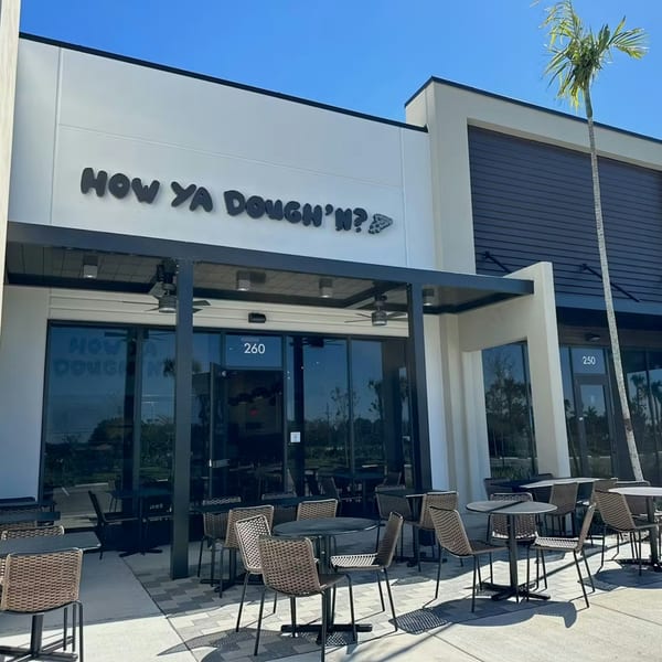 The exterior of How Ya Dough'n Pizza, featuring outdoor seating, large windows, and a palm tree.